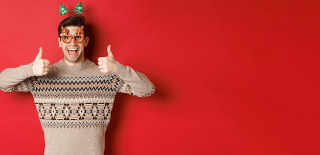 Retrato de un hombre satisfecho y feliz con suéter navideño y gafas de fiesta mostrando el pulgar hacia arriba deseando feliz año nuevo parado sobre fondo rojo