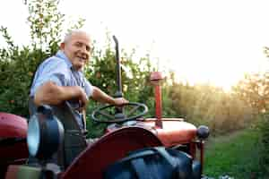 Foto gratuita retrato de hombre senior agricultor conduciendo su vieja máquina tractor de estilo retro a través de huerto de manzanas en la puesta del sol