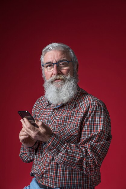 Retrato de un hombre senior con lentes sosteniendo un teléfono inteligente en la mano contra el telón de fondo rojo