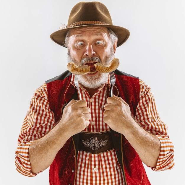 Retrato de hombre senior de Oktoberfest con sombrero, vistiendo la ropa tradicional bávara. Tiro de cuerpo entero masculino en estudio sobre fondo blanco. La celebración, vacaciones, concepto de festival. Comiendo salchichas.