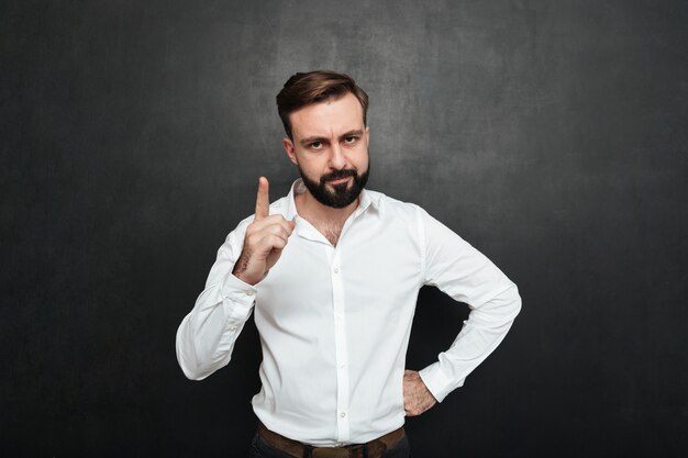 Retrato de hombre serio 30s en camisa blanca posando en la cámara con mostrar el dedo hacia arriba aislado sobre gris oscuro