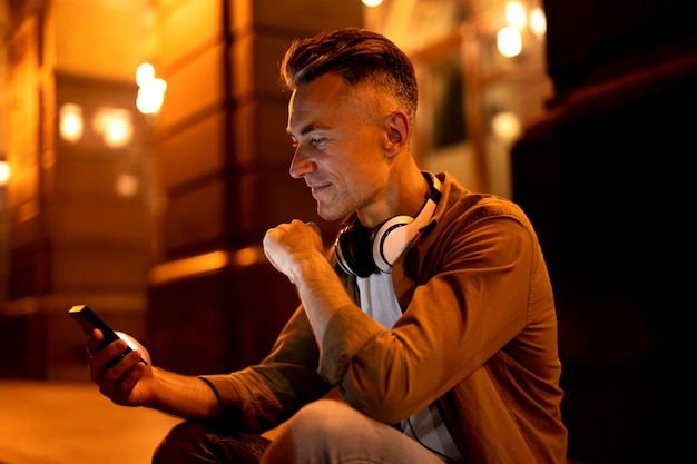 Retrato de hombre sonriente en la ciudad por la noche con auriculares y smartphone