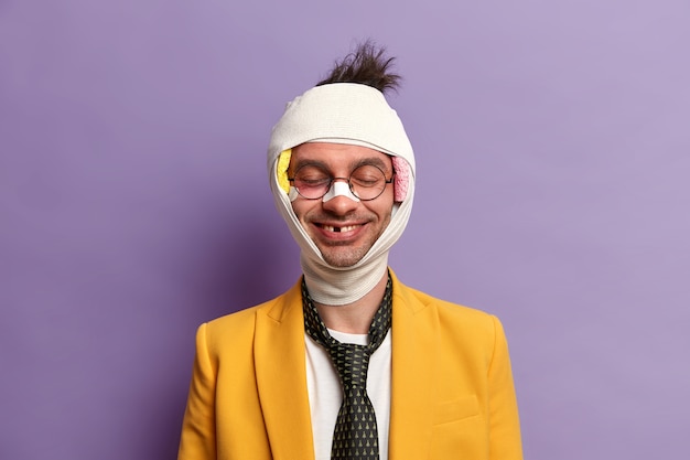 Foto gratuita retrato de hombre sonriente divertido al que le faltan dientes después de un trauma grave, se para con los ojos cerrados, piel magullada, cabeza vendada, se cayó durante la bicicleta, tiene un período de recuperación, aislado en una pared púrpura