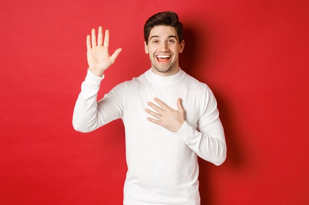 Retrato de hombre sonriente honesto en suéter blanco haciendo una promesa jurando decir la verdad de pie aga ...