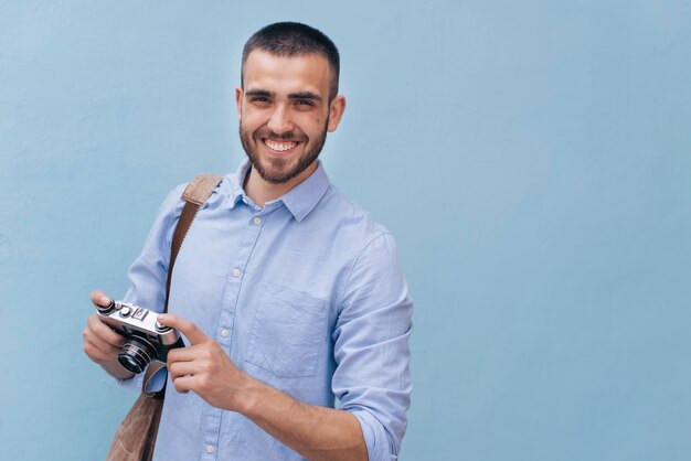 Retrato del hombre sonriente joven que sostiene la cámara que se opone a la pared azul