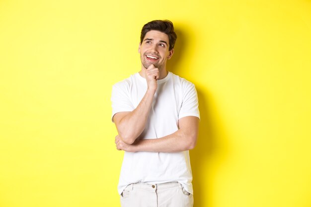 Retrato de hombre sonriente mirando pensativo en la esquina superior izquierda, elegir algo, tener una idea, de pie contra el fondo amarillo.