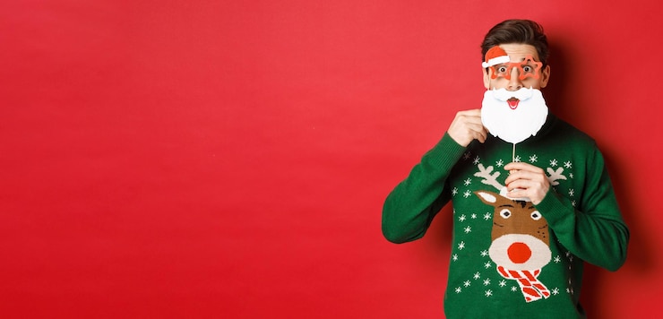 Foto gratuito retrato de un hombre sorprendido con un suéter verde de navidad sosteniendo una divertida máscara de papá noel celebrando la nueva...