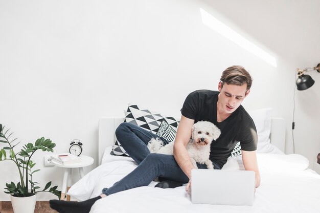 Retrato de un hombre con su caniche de juguete usando la computadora portátil en el dormitorio