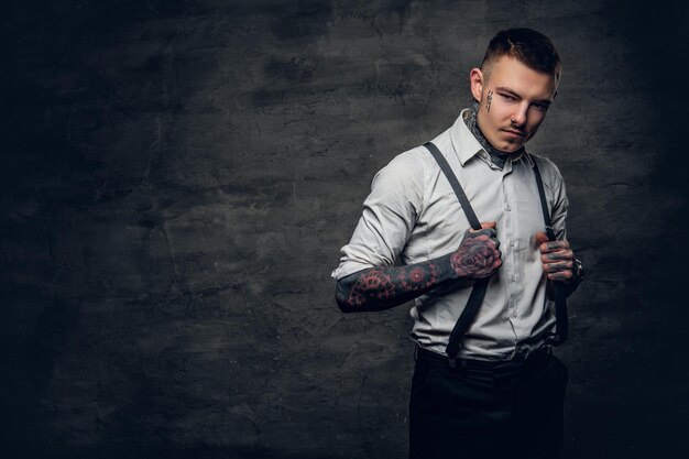 Retrato de un hombre con un tatuaje en la cara y los brazos, vestido con una camisa blanca sobre un fondo gris.