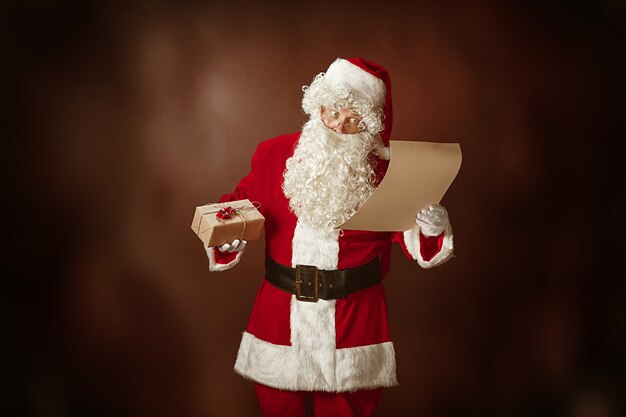 Retrato de hombre en traje de Santa Claus - con una lujosa barba blanca, sombrero de Santa y una carta de lectura de traje rojo en el fondo rojo del estudio con regalos