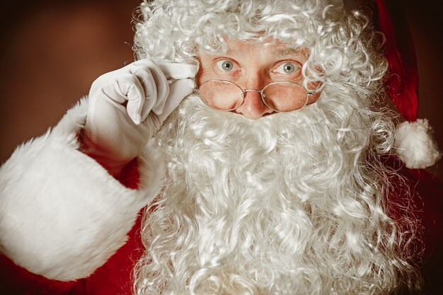 Retrato de hombre en traje de Santa Claus - con una lujosa barba blanca, sombrero de Santa y un traje rojo en el fondo rojo del estudio. La cara de cerca