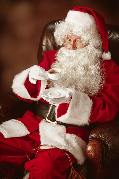 Retrato de hombre en traje de Santa Claus - con una lujosa barba blanca, sombrero de Santa y un traje rojo en el fondo rojo del estudio sentado en una silla con una taza de café