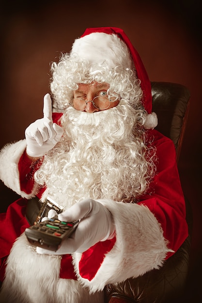 Retrato de hombre en traje de Santa Claus con una lujosa barba blanca, sombrero de Santa y un traje rojo en rojo sentado en una silla con control remoto de TV