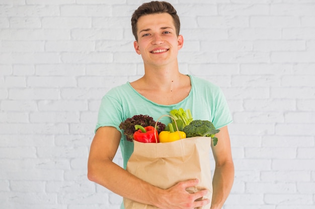 Retrato de hombre con verduras frescas y frutas en bolsa marrón contra la pared de ladrillo