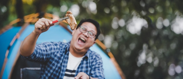 Retrato de hombre viajero asiático anteojos bistec de cerdo freír barbacoa en una sartén para asar o una olla en un camping Cocina al aire libre viajar concepto de estilo de vida de camping