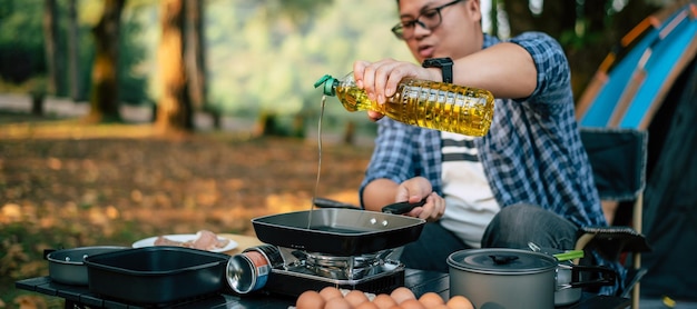 Retrato de hombre viajero asiático anteojos vertiendo aceite de girasol en una sartén Cocina al aire libre viajar camping concepto de estilo de vida