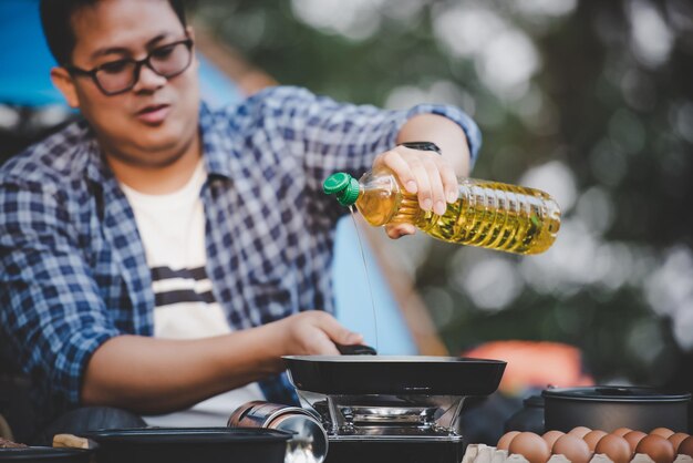 Retrato hombre viajero tailandés gafas verter aceite de girasol en una sartén cocina al aire libre viajar camping concepto de estilo de vida