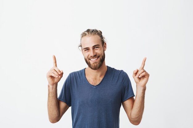 Retrato de hombres hermosos alegres con peinado de moda y barba en camiseta azul riendo y apuntando hacia arriba