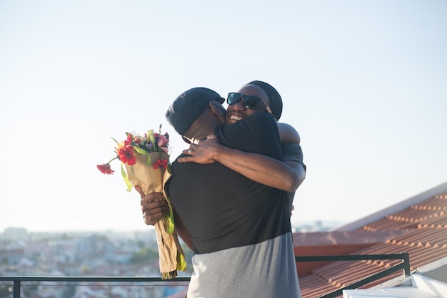 Retrato de hombres homosexuales afroamericanos abrazándose en la azotea. Dos hombres felices parados en el fondo del cielo azul abrazándose unos a otros un hombre sosteniendo un ramo de flores. Relaciones de pareja LGBT, concepto de igualdad
