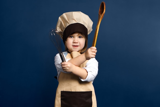 Retrato horizontal de adorable niña caucásica en uniforme de chef cruzando los brazos sosteniendo una cuchara de madera y un batidor, posando sobre fondo de pared de estudio en blanco con espacio de copia para su contenido