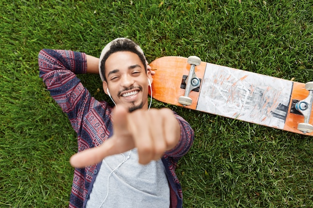 Retrato horizontal del alegre patinador masculino barbudo se encuentra en la hierba verde cerca de la patineta, escucha música con auriculares