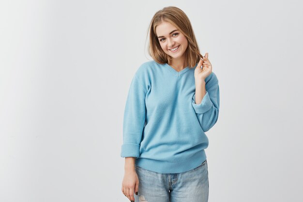 Retrato horizontal de atractiva mujer de ojos oscuros vestida casualmente con aspecto juguetón mientras sonríe. Mujer joven hermosa que juega con su pelo mientras que presenta en el estudio.