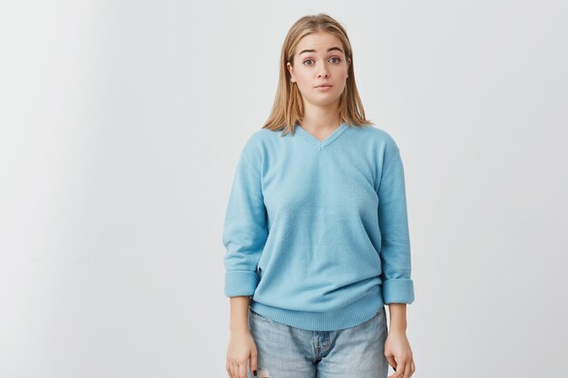 Retrato horizontal de una atractiva mujer de ojos oscuros vestida con un suéter azul y jeans que parece sorprendida y conmocionada por las noticias que acaba de escuchar. Chica guapa posando.