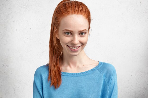 Retrato horizontal de feliz modelo de mujer joven con expresión alegre, tiene cola de caballo roja brillante, vestida de manera informal, sonríe agradablemente, se regocija por recibir elogios del hombre