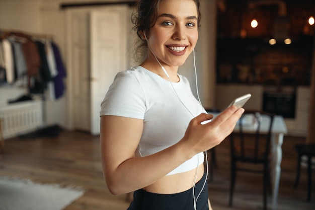 Retrato horizontal de hermosa deportista elegante en entrenamiento superior recortado en interiores con auriculares, usando el teléfono celular, escuchando pistas de música dinámicas