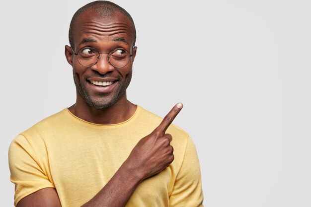 Retrato horizontal de hombre feliz mira alegremente a un lado, señala con el dedo índice en la esquina superior derecha