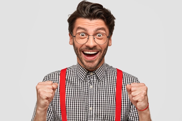 El retrato horizontal de un joven europeo alegre tiene expresión facial llena de alegría, aprieta los puños con entusiasmo, usa camisa a cuadros y tirantes rojos, expresa felicidad después de ganar el concurso