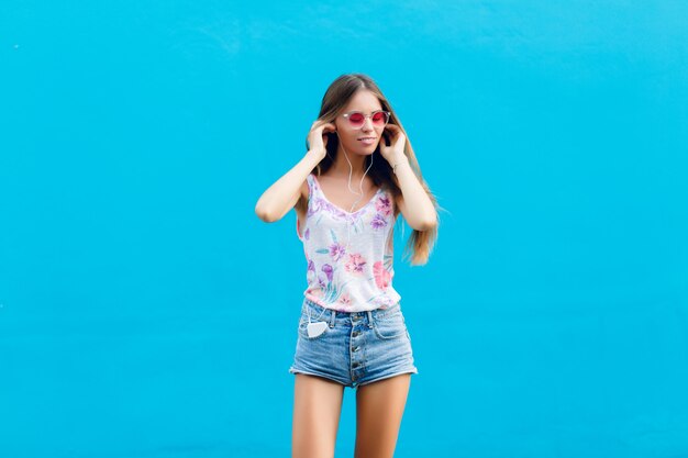 Retrato horizontal de linda chica elegante sobre fondo azul está parado y escucha música en auriculares en el teléfono inteligente