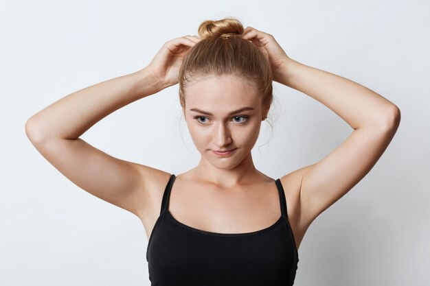 Retrato horizontal de mujer atractiva con nudo de cabello rubio, mirando pensativamente hacia abajo con sus ojos azules mientras intenta hacer moño, pensando en dónde ir a caminar con amigos. Concepto de belleza