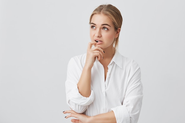 Retrato horizontal de mujer confundida con el pelo liso teñido de rubio, sosteniendo su dedo sobre los dientes haciendo difícil elegir no saber qué elegir.