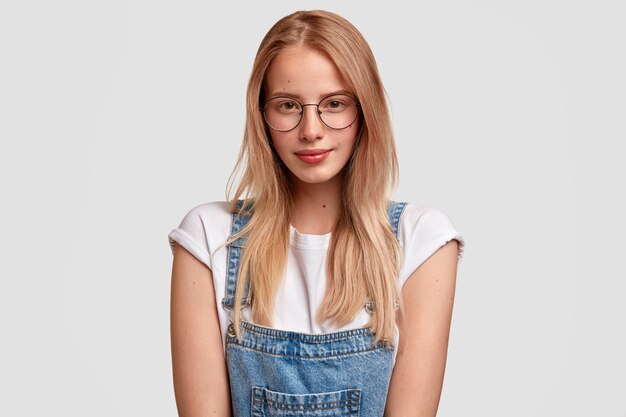 Retrato horizontal de mujer mujer de aspecto agradable en gafas y overoles de mezclilla, listo para escuchar conferencias, se prepara para las clases, está parado contra la pared blanca. Concepto de personas, juventud y estilo.