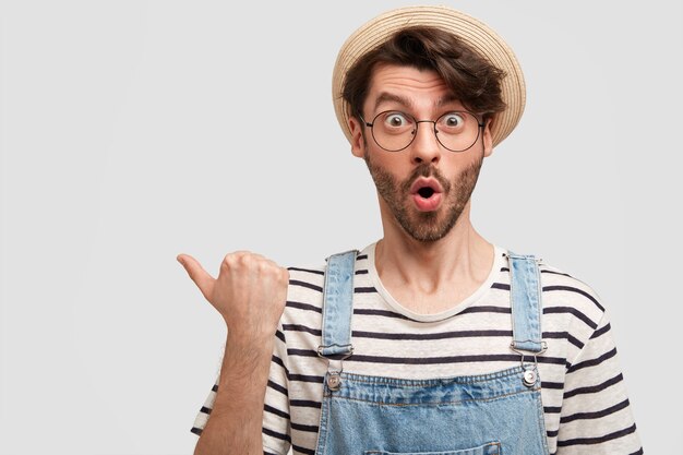 El retrato horizontal de un trabajador agrícola de sexo masculino hermoso y estupefacto mantiene la boca abierta, señala con el pulgar a la izquierda, muestra un espacio en blanco, nota algo fantástico, aislado sobre una pared blanca