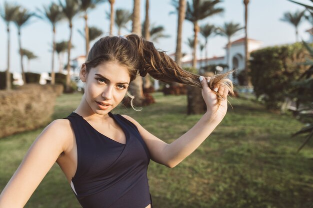 Retrato increíble joven atractiva en ropa deportiva jugando con su pelo largo y rizado en el parque de la ciudad tropical. Mirando, modelo delgado de moda, resort, juguetón, outwork