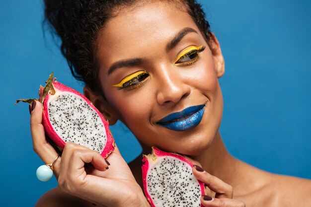 Retrato de una increíble mujer mulata con maquillaje brillante disfrutando de exóticas frutas pitaya cortadas por la mitad y mirando a un lado aislado, sobre la pared azul