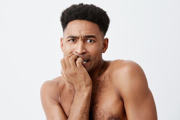 Retrato de infeliz hombre africano de piel oscura con el pelo rizado y el cuerpo desnudo con la mano cerca de la boca, mirando a un lado con expresión de la cara culpable y estresada.