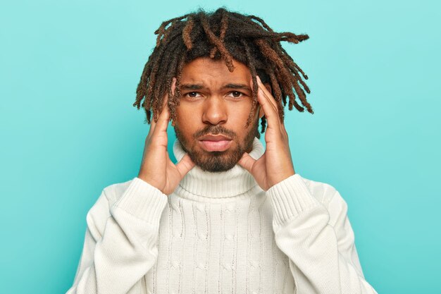 Retrato de infeliz hombre de piel oscura con rastas, sufre de dolor de cabeza insoportable, mantiene las manos en las sienes, viste suéter blanco con cuello, aislado sobre fondo azul, pide analgésicos