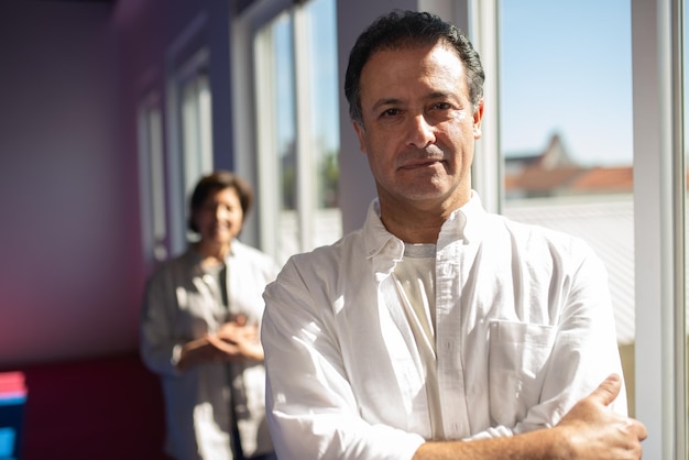 Retrato de un instructor de yoga serio y confiado con camisa blanca parado contra un estudiante en el gimnasio con ventanas panorámicas. concepto de entrenador