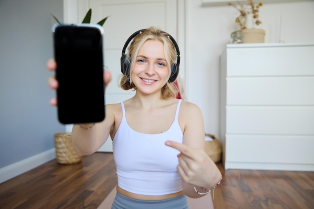 Foto gratuita retrato de una instructora de fitness mostrando su aplicación de entrenamiento favorita apuntando con el dedo al teléfono inteligente