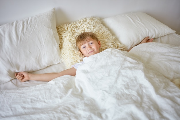 Retrato de interior de un adolescente europeo soñoliento estirando los brazos después de despertarse temprano en la mañana, acostado sobre sábanas blancas, yendo a la escuela, mirando, con expresión facial perezosa