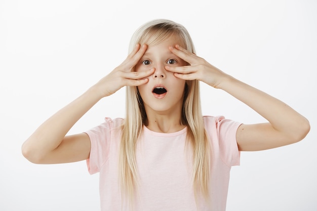 Retrato interior de una adorable niña rubia sorprendida sorprendida con una camiseta rosa, dejando caer la mandíbula, diciendo wow mientras mantiene los dedos cerca de los ojos