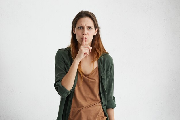 Retrato interior aislado de una mujer europea joven enojada enojada en ropa casual frunciendo el ceño, gesticulando con el dedo, manteniéndolo en los labios, pidiendo guardar silencio