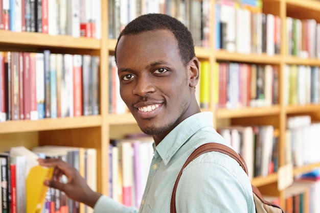 Retrato interior del apuesto hombre africano recogiendo libro de estantería en la librería. Estudiante feliz negro pasando vacaciones en la biblioteca de la universidad, tomando prestado un libro de texto para investigación