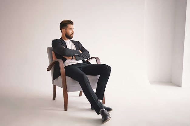 Retrato interior de un apuesto joven abogado con barba espesa y peinado moderno sentado cómodamente en un sillón, manteniendo los brazos cruzados sobre el pecho y mirando hacia otro lado con expresión pensativa