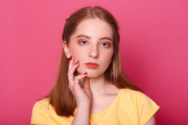 Retrato interior de una atractiva chica guapa con el pelo largo y liso, tiene una expresión facial seria, mantiene la mano en la mejilla