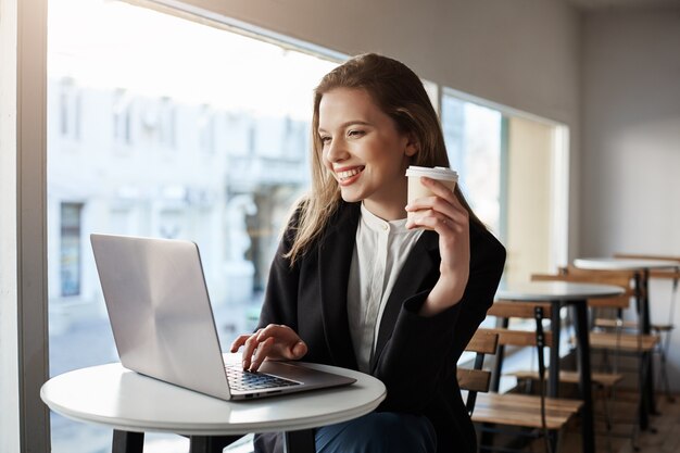 Retrato interior de atractiva mujer europea sentada en la cafetería, tomando café y escribiendo en la computadora portátil, ser feliz y satisfecho.