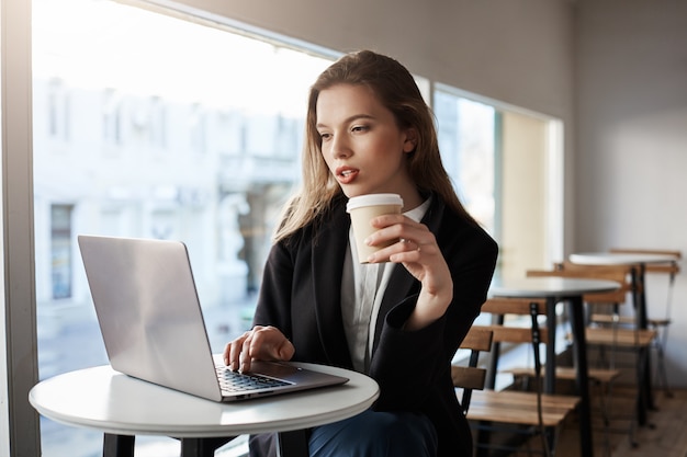 Retrato interior de atractiva mujer europea sentada en la cafetería, tomando café y escribiendo en la computadora portátil